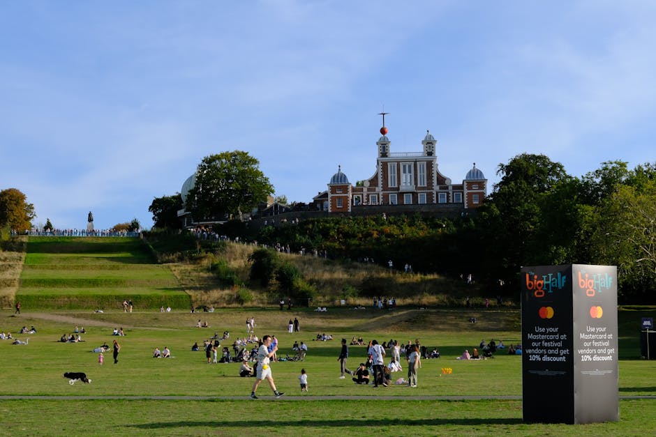 A historic building with a domed roof and classical architectural details is situated in the foreground, surrounded by a wrought iron fence and a grassy area with a few benches and people walking near