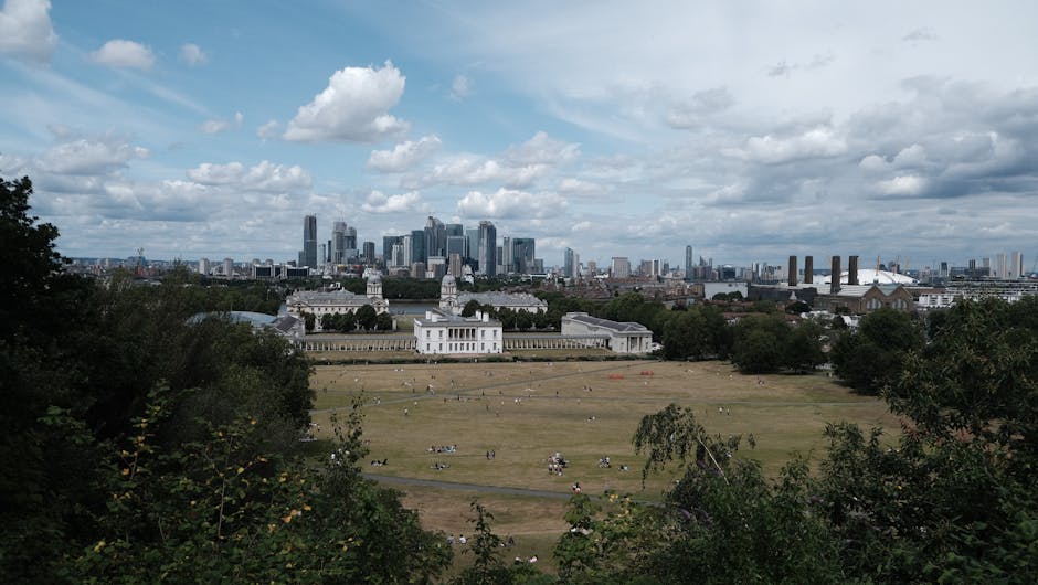 A wide view of the historic Old Royal Naval College buildings with their domed roofs and classical architecture, situated in Greenwich, London, with modern high-rise office buildings visible in the ba