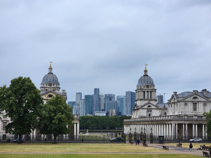 A wide-angle view of a park with a gravel pathway leading towards two symmetrical historic white stone buildings with domed towers and classical architectural features, situated on either side of the 