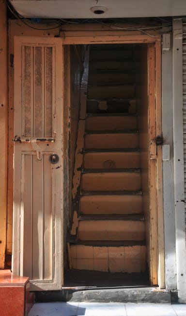 An interior view of a narrow staircase within an apartment or house, featuring wooden steps with a dark finish and a metal handrail along the side. To the right, there is a tall, narrow window allowin